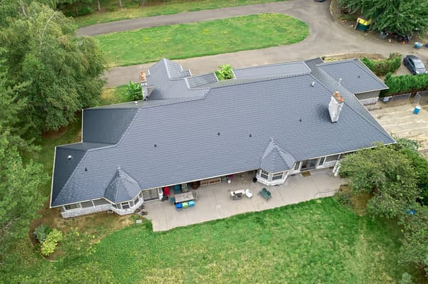 Aerial rear view of a large ranch-style home in Ridgefield, WA with a complex Interlock aluminum slate roof in charcoal black, featuring turreted dormers and two chimneys.
