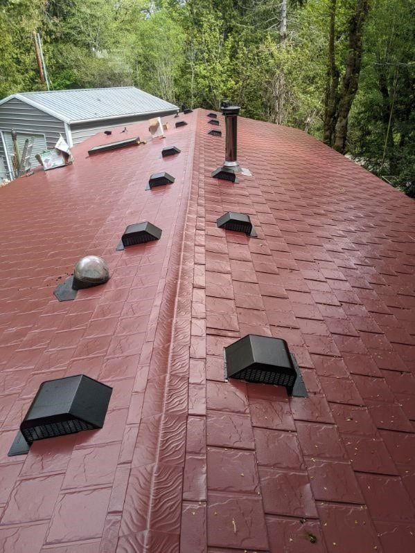 Aerial view of a tile-red Interlock aluminum slate metal roof on a single-family home, featuring multiple roof louvers, a vent pipe, and chimney, surrounded by forest.
