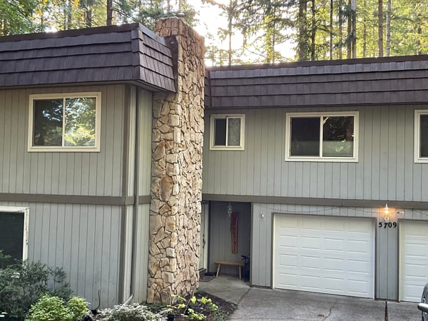 Two-story home in Olympia, WA with Interlock aluminum shake roofing in Colonial Brown, prominent stone chimney, and wooded backdrop.