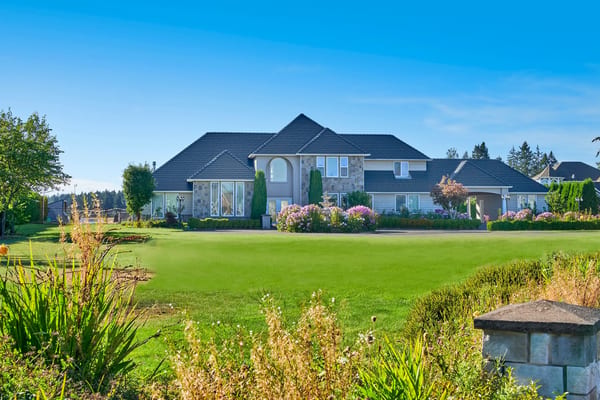 Large two-story suburban home with deep charcoal multi-hip metal cedar shingle roof, stone accents, manicured lawn, and blooming landscaping.