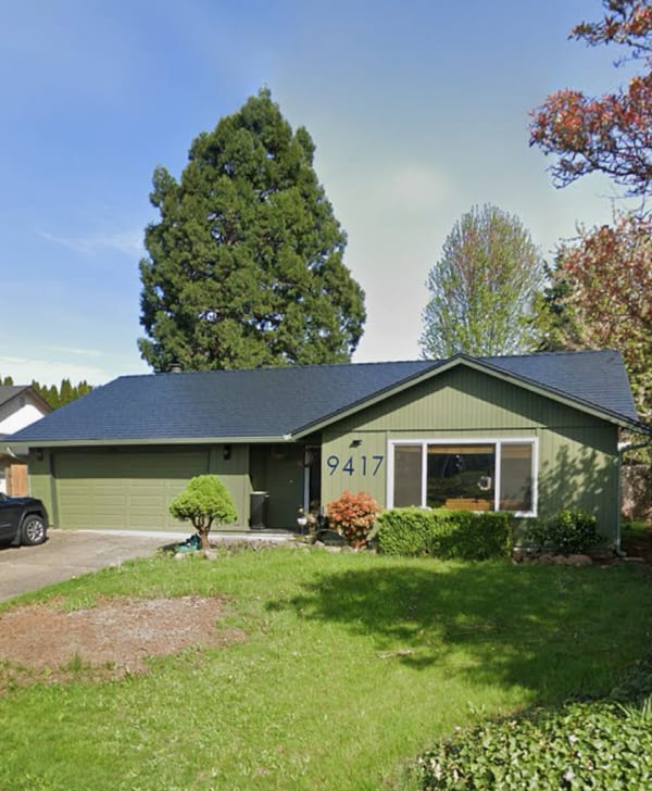 Single-story ranch home with black Interlock aluminum cedar shingle metal roof, green siding, attached two-car garage, and mature evergreen tree backdrop.