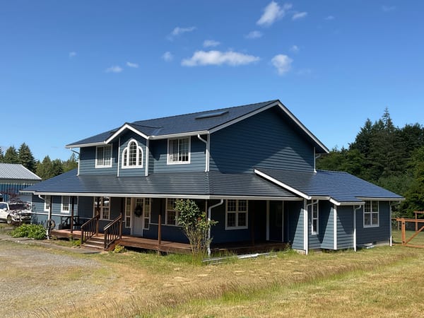 Two-story farmhouse with deep charcoal Interlock aluminum cedar shingle metal roof, wrap-around porch, dormer, and skylights in rural Olympia, WA.