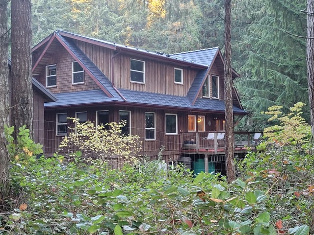 Two-storey cedar-clad chalet nestled in forest with a complex steep deep charcoal Interlock aluminum slate metal roof featuring multiple dormers and valleys.