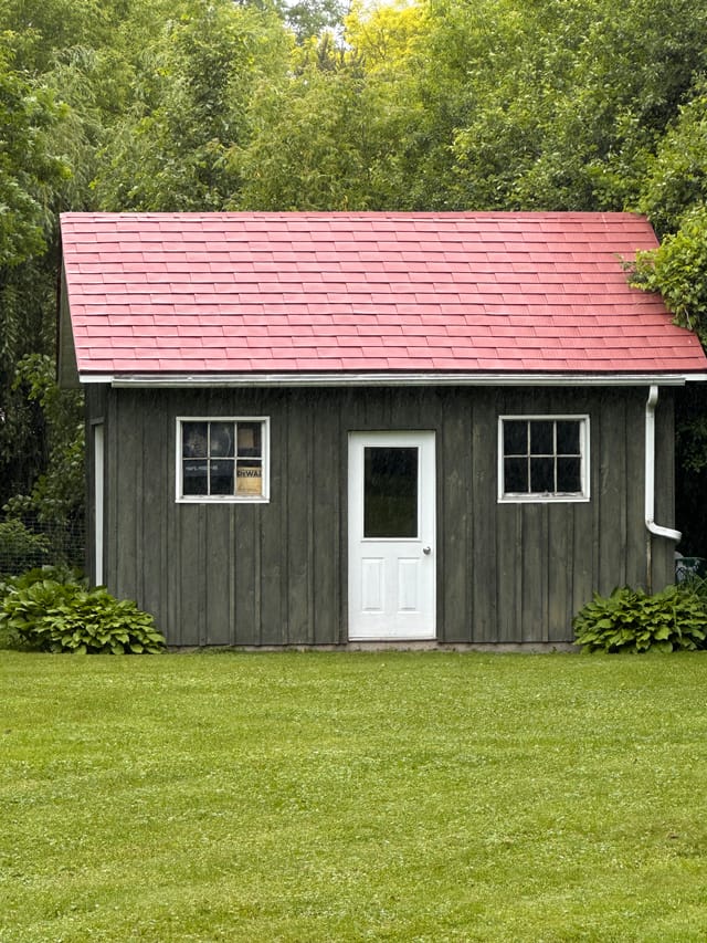 Front elevation of a rustic wood-clad outbuilding with a Tile Red Interlock aluminum shingle gable roof, white door, and twin windows in Almonte, ON.