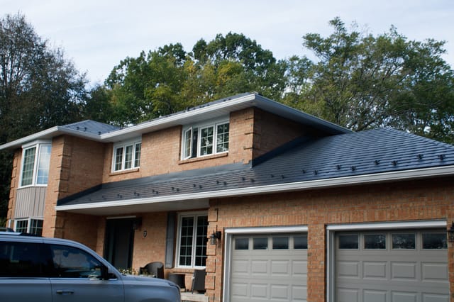 Two-storey brick colonial home with deep charcoal Interlock aluminum slate metal roof, snow guards visible, set against a forested suburban backdrop.