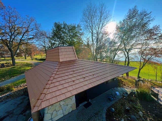 Aerial view of a modern single-family home with Interlock Copper Penny aluminum shake metal roofing, multi-hip design, and matching copper chimney wrap.