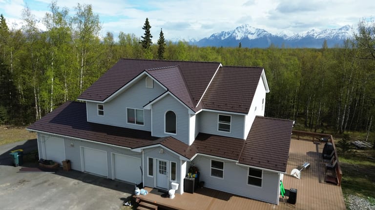 A two-story house with a brown metal roof and white siding, set amidst lush greenery and a mountain backdrop.