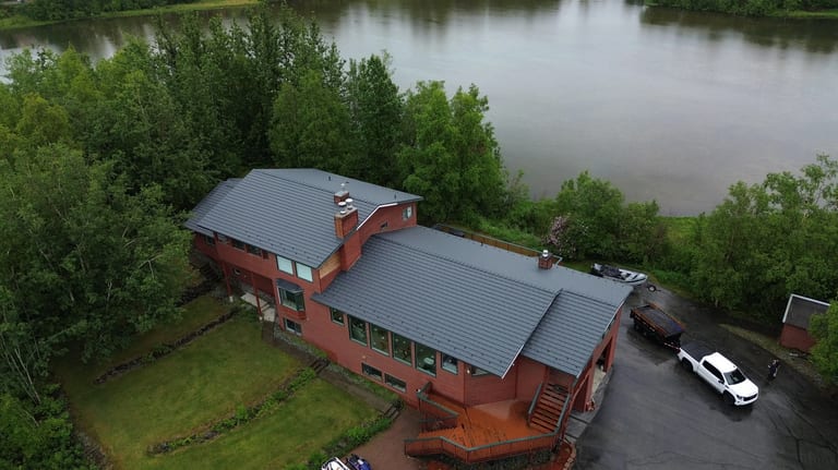 A modern single-family home with a metal roof and a chimney, overlooking a waterfront and forested area on a lush greenery day with overcast sky.