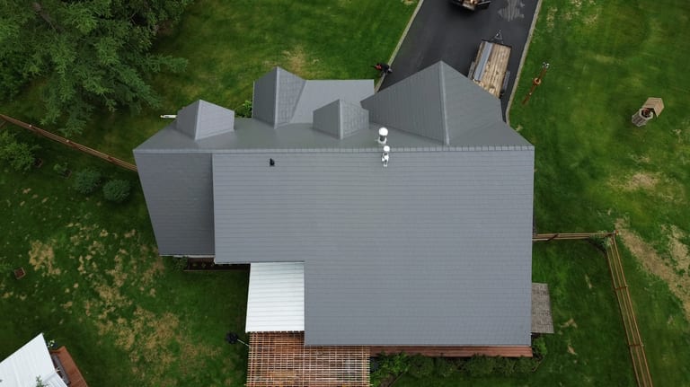 Aerial view of a modern, single-family home with a grey metal roof, snow guards and rails, and a roof louver, in a rural setting with lush greenery.