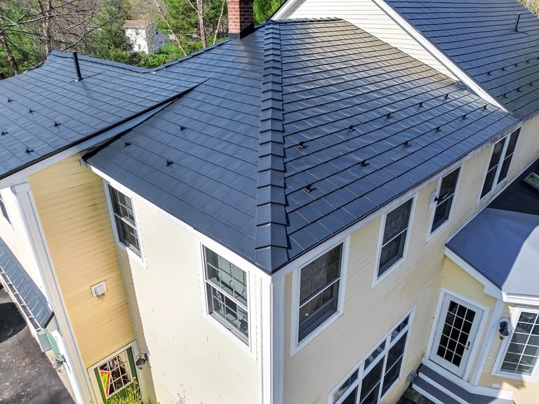 Black Interlock Slate aluminum roof on a yellow two-storey home with dormers among tall pines, Brunswick, Maine