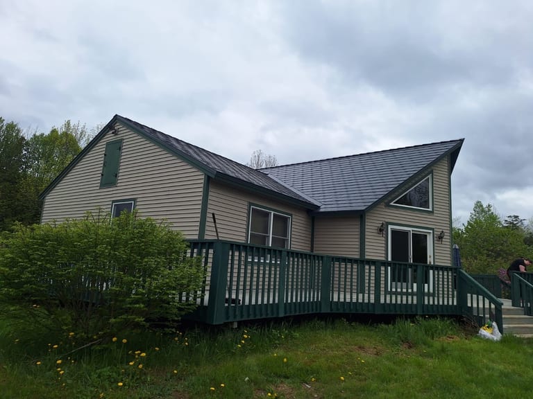 1.5-story rural farmhouse with Interlock deep charcoal aluminum slate metal roof, complex multi-plane gable design, tan vinyl siding, and dark green deck in Stetson, ME.
