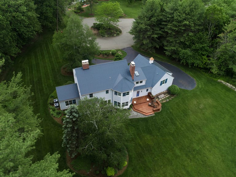 Aerial rear view of a large Colonial home in Topsfield, MA with a complex deep charcoal Interlock aluminum slate metal roof, two chimneys, and a rear wood deck.