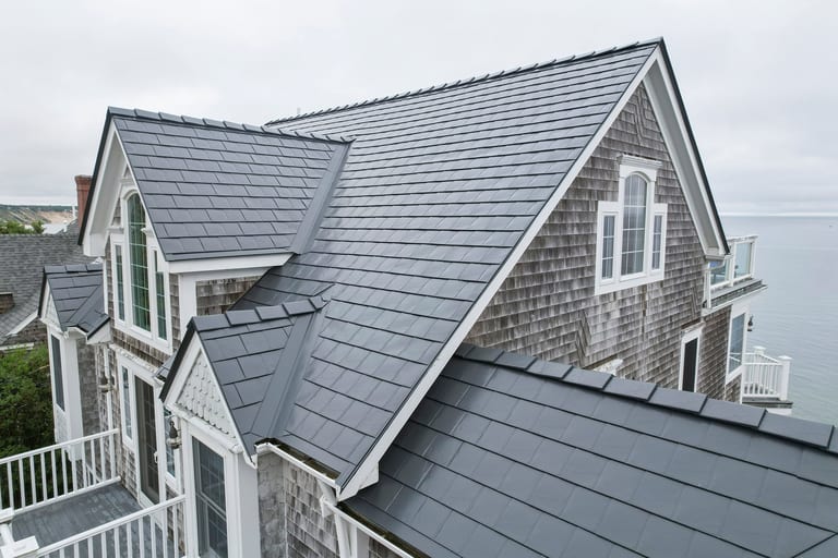 Aerial front view of a coastal shingle-sided home with a complex steep Interlock aluminum slate roof in Deep Charcoal, featuring multiple dormers and valleys.