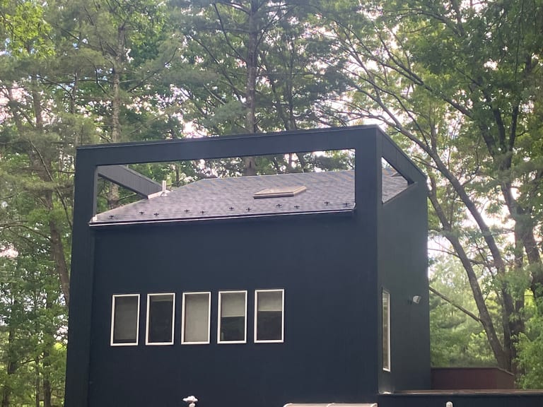 Modern black two-story home with Interlock deep charcoal aluminum slate roof, skylight, and vent pipe visible above a row of horizontal windows, surrounded by forest.
