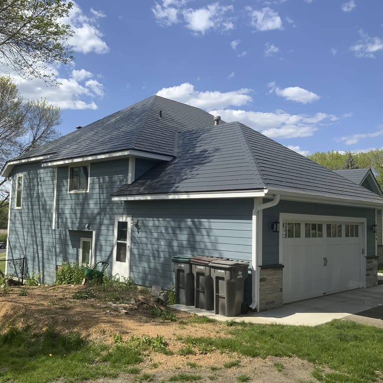Two-story suburban home with charcoal grey Interlock aluminum slate metal roofing on a complex multi-hip profile, blue lap siding, and attached garage.
