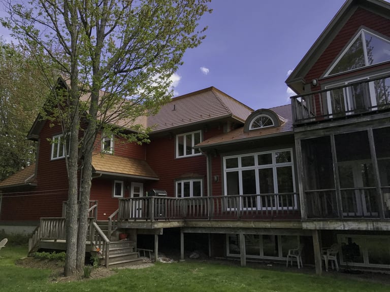 Aged Copper Interlock Cedar Shingle aluminum roof on a red two-storey home with a metal roof among tall pines, Hanwell, New Brunswick