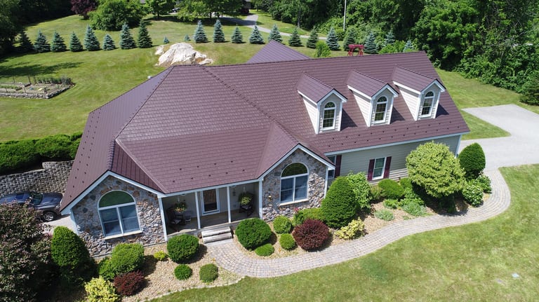 Aerial front view of a 1.5-story Colonial home in NH featuring an Interlock Colonial Brown cedar shingle metal roof with dormers and stone facade.