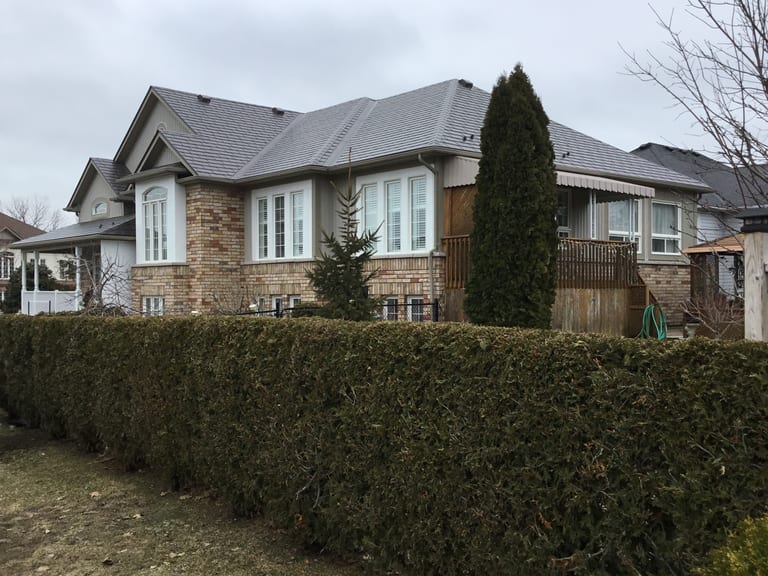 Large single-family home with complex multi-hip Interlock aluminum slate roof in Tudor Brown, brick exterior, viewed from front-right across a trimmed cedar hedge.