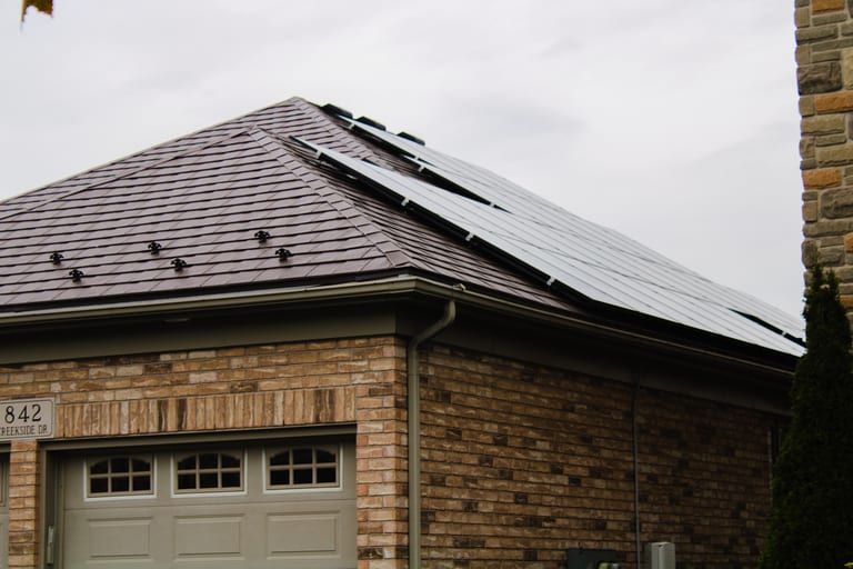 Colonial Brown Interlock aluminum slate metal roof on a brick suburban home with solar panels, snow guards, and hip-style multi-plane roofline.
