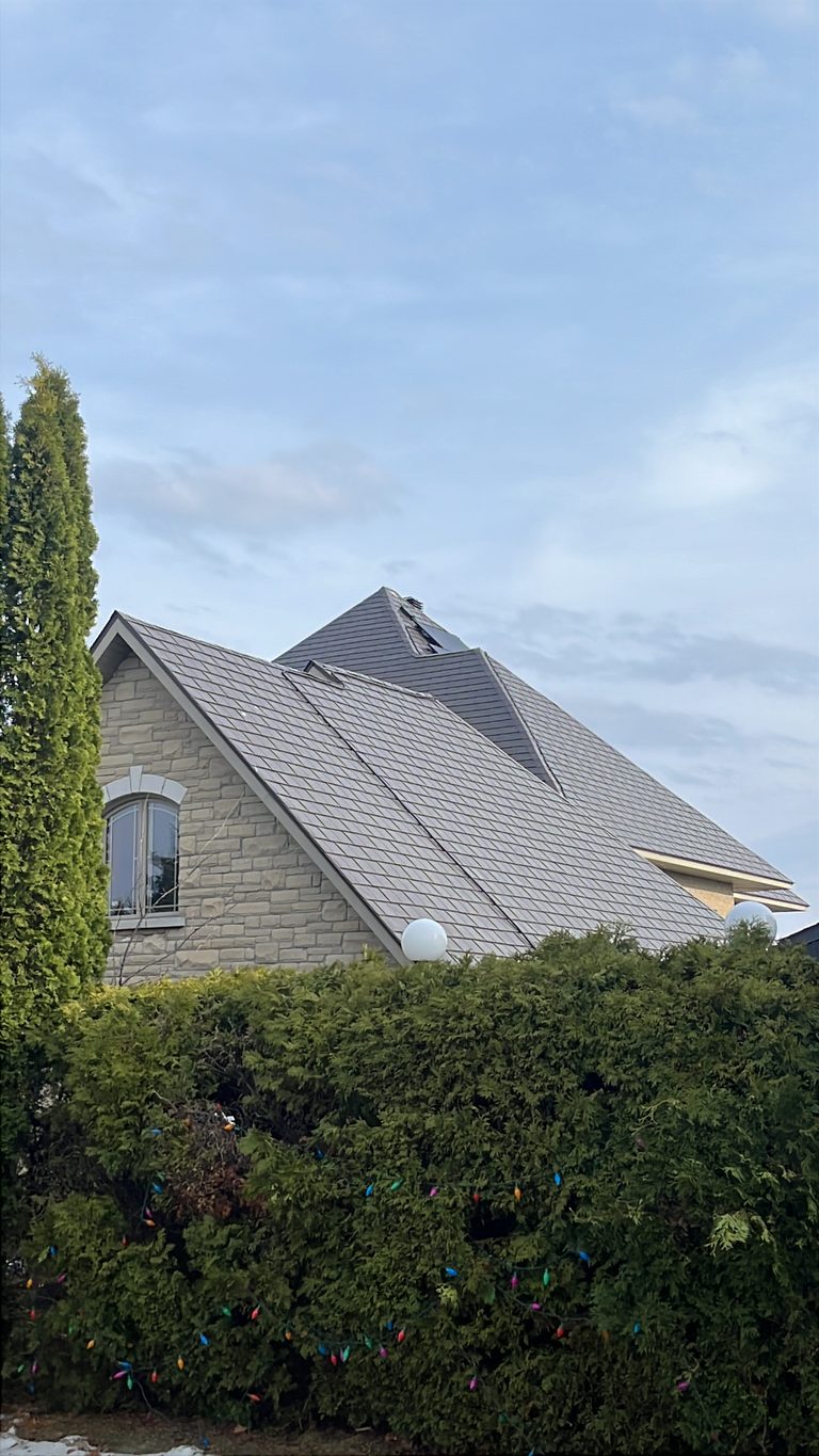 Steep multi-hip Interlock aluminum slate roof in Tudor Brown on a stone two-storey home in Sudbury, ON, framed by tall cedars and holiday lights.