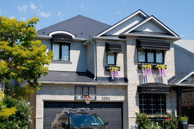 Two-storey suburban home in Milton ON with black Interlock aluminum slate metal roofing, arched dormer, dark awnings, and light brick exterior.