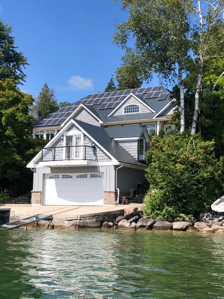Craftsman lakefront home with deep charcoal Interlock aluminum slate roof, multiple solar panels, complex multi-gable profile, and boathouse garage on rocky shoreline.