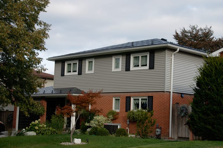 Two-storey suburban home with black Interlock aluminum slate metal roofing, red brick lower facade, grey vinyl siding, and multi-hip roof profile in Scarborough, ON.