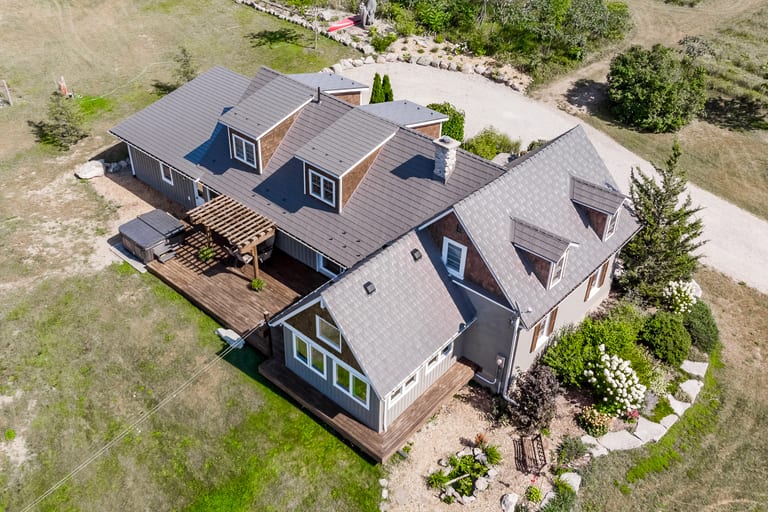 Aerial rear view of a 1.5-storey rural home in Midland, ON with Interlock weathered cedar shingle metal roofing, multiple dormers, chimney, and wood deck.