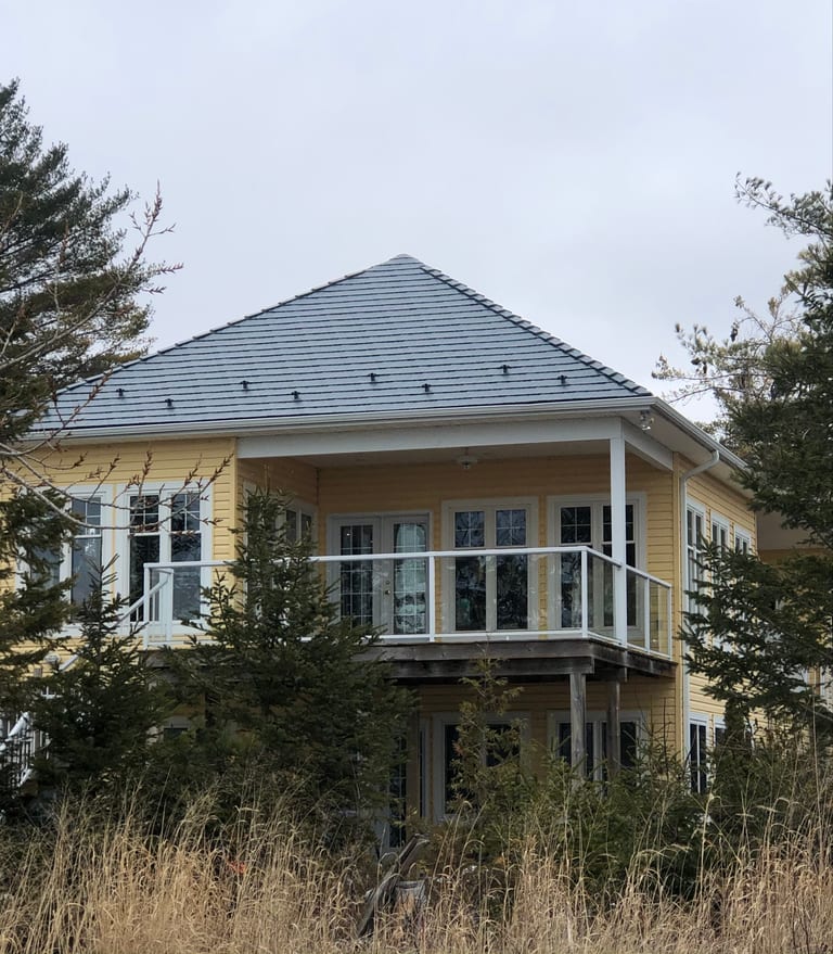 Two-storey yellow cottage with a hip-profile deep charcoal Interlock cedar shingle metal roof, snow guards, and a covered upper deck balcony, set among coastal grasses and pines.