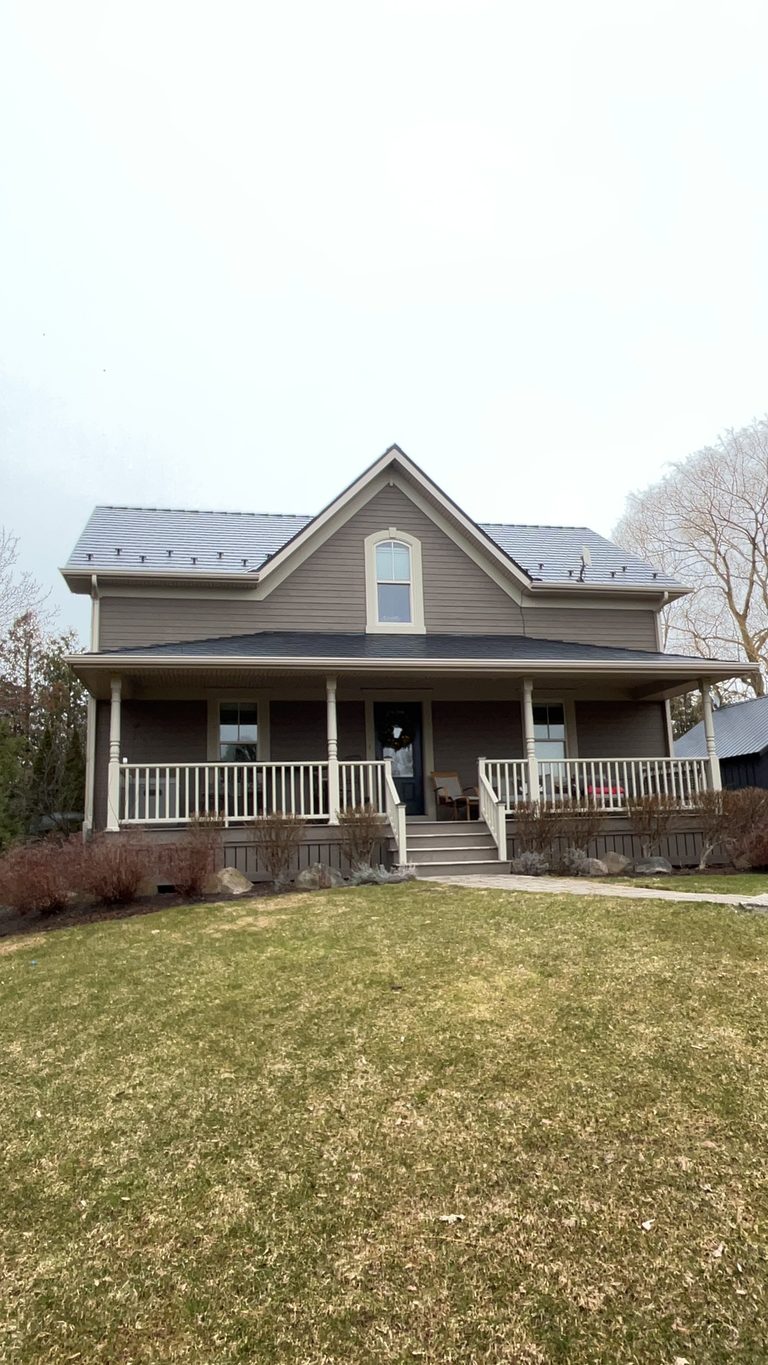 Two-storey farmhouse in Ravenna, ON with black Interlock aluminum slate metal roof, front dormer, wrap-around porch, and snow guards visible on steep gable.