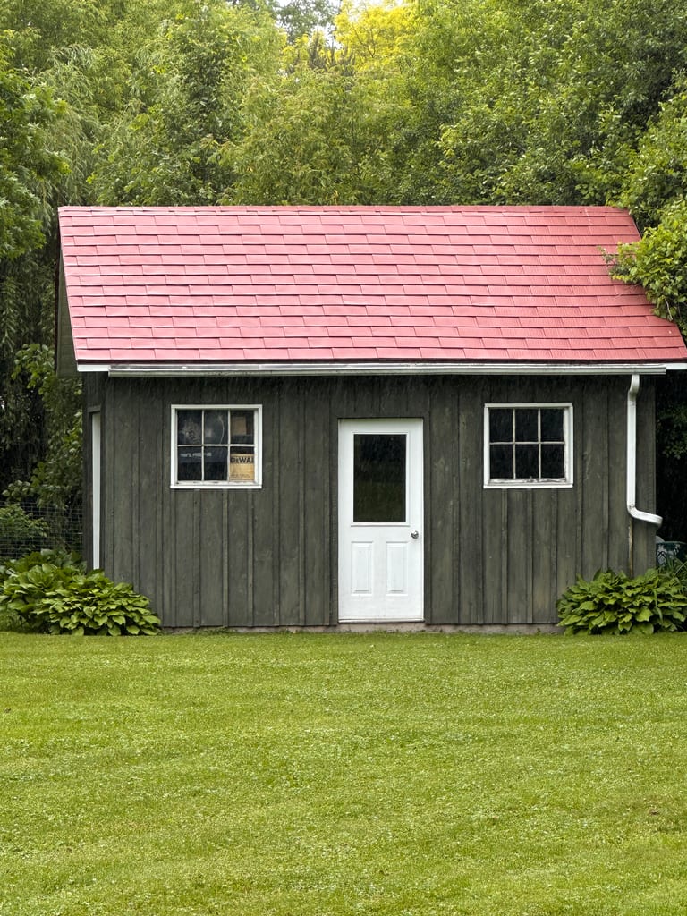 Front elevation of a rustic wood-clad outbuilding with a Tile Red Interlock aluminum shingle gable roof, white door, and twin windows in Almonte, ON.