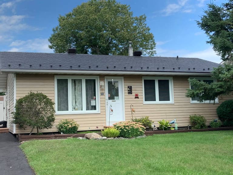 Single-story ranch home in Ottawa with Interlock black aluminum slate metal roof, low-pitch simple gable, tan vinyl siding, and manicured front garden.