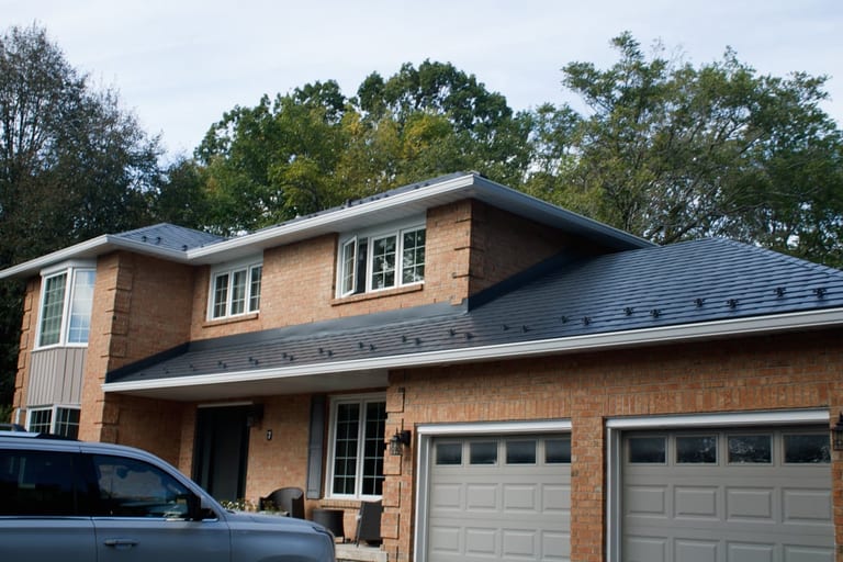 Two-storey brick colonial home with deep charcoal Interlock aluminum slate metal roof, snow guards visible, set against a forested suburban backdrop.