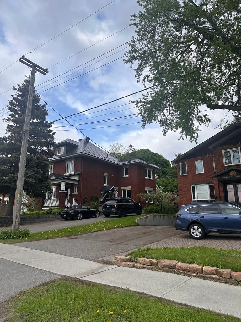 Street-view of a red-brick heritage home in Ottawa with a steep black Interlock aluminum slate metal roof, multi-hip profile, surrounded by mature trees.