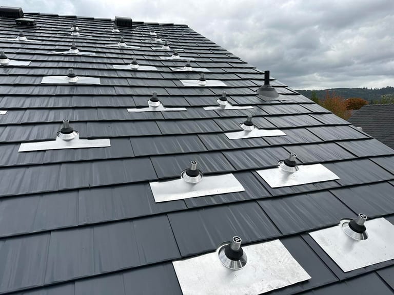 Close-up of deep charcoal Interlock aluminum cedar shingle metal roof with multiple aluminum-flashed vent pipe penetrations on a hip roof.