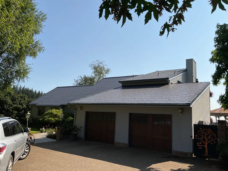 After: Deep Charcoal Interlock Slate aluminum roof on a beige ranch home with a brown garage door among tall trees, Amity, Oregon