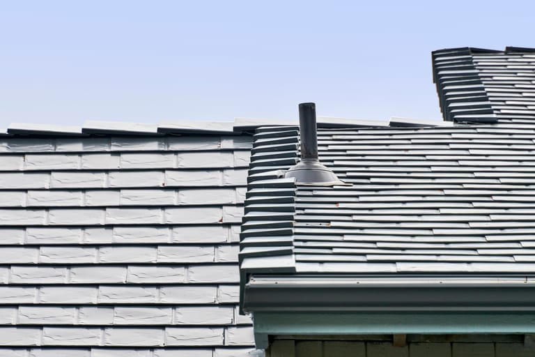 Close-up of Interlock deep charcoal aluminum slate roofing showing multi-level roof transition with vent pipe flashing detail against a clear blue sky.
