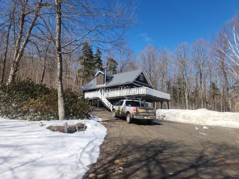 Chalet-style Vermont home with Interlock Musket Grey cedar shingle metal roof, steep gable profile, and white wraparound deck, surrounded by bare winter trees.