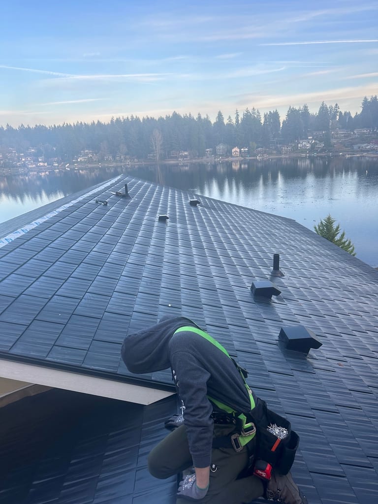 Roofer in safety harness installs black Interlock aluminum cedar shingle roofing on a lakeside home in Lakewood, WA, with calm water and trees behind.