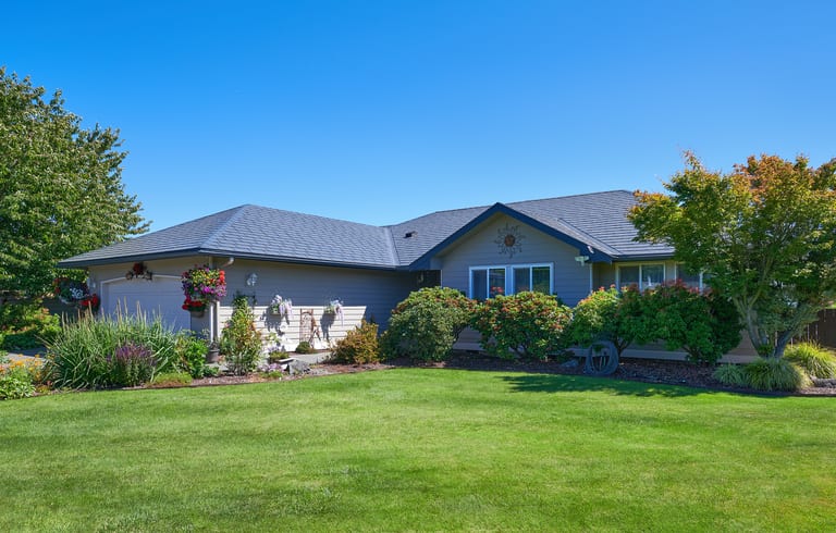 Single-story ranch home with Interlock deep charcoal aluminum slate roof featuring multi-hip complexity, lush manicured landscaping, and attached garage in Sequim, WA.