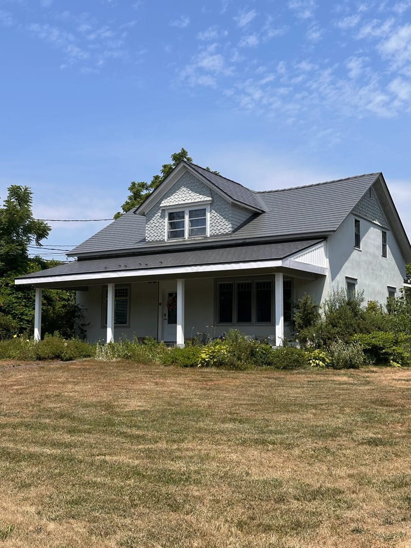 Musket Grey Interlock Slate aluminum roof on a grey two-storey home with dormers among tall pines, Bloomfield, Ontario