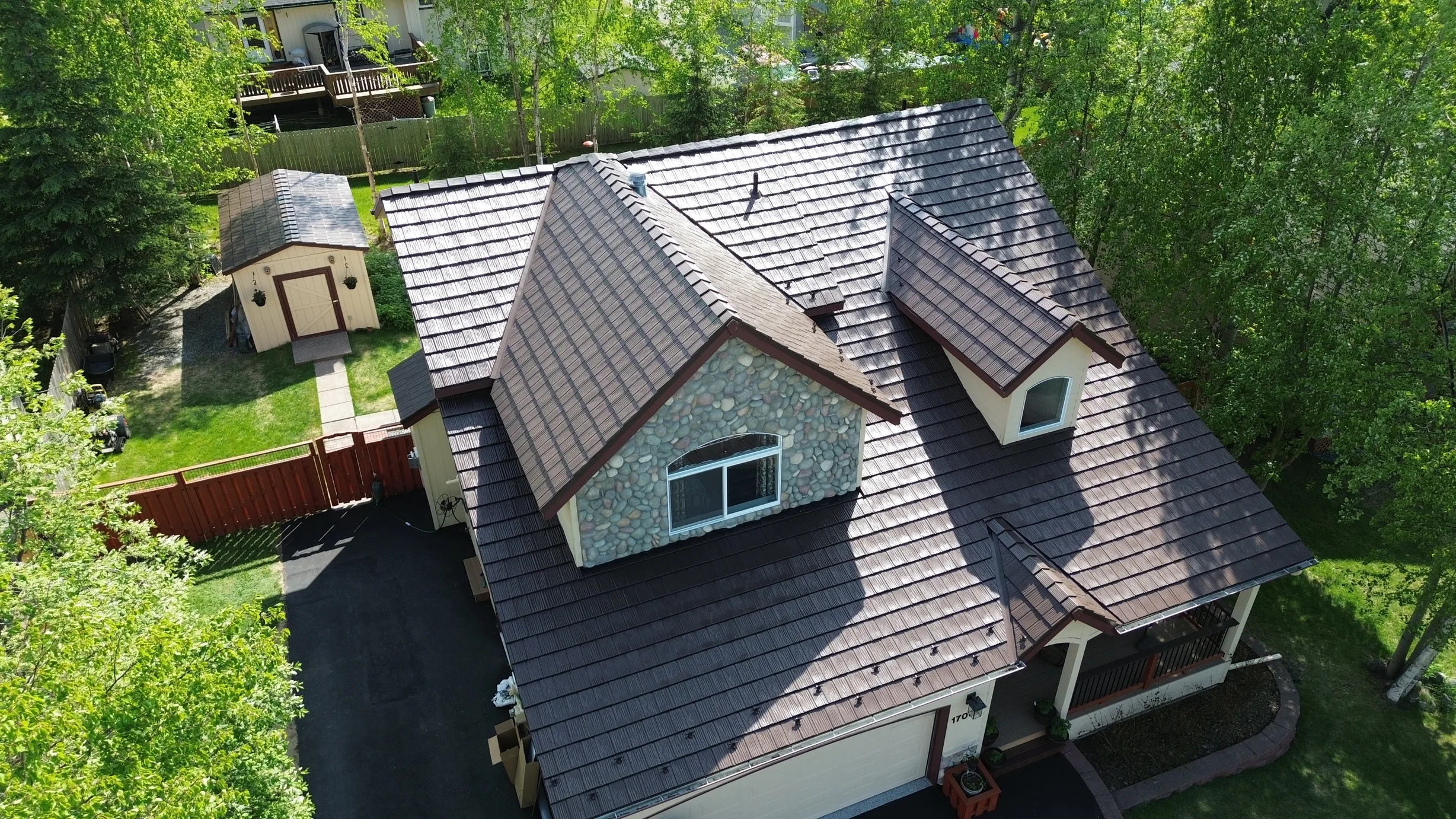 A brown metal roof on a white cottage with brown trim, set amidst lush greenery.