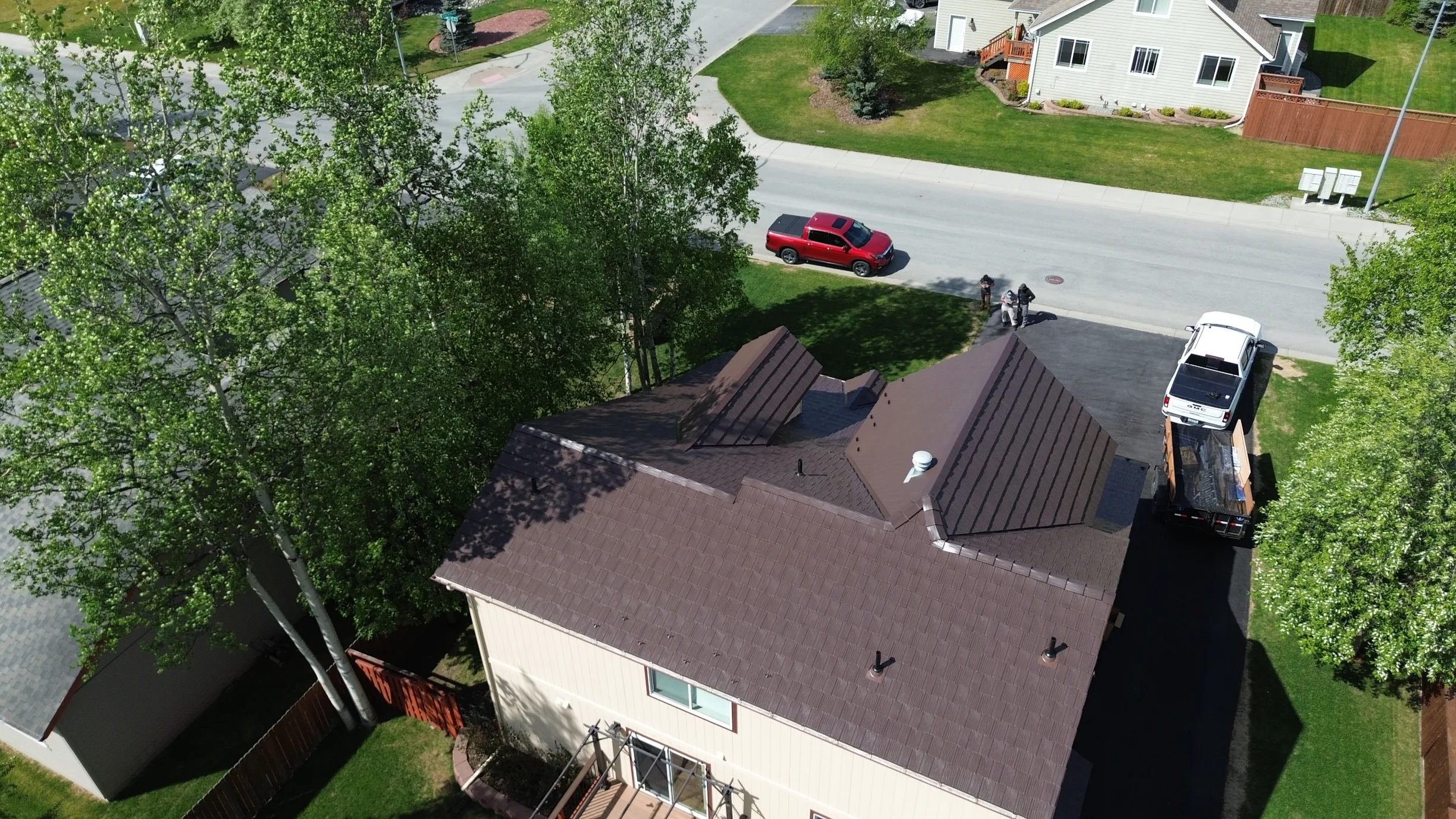 A modern single-family home with a brown metal roof, viewed from above, in a rural or suburban setting with lush greenery and blue sky