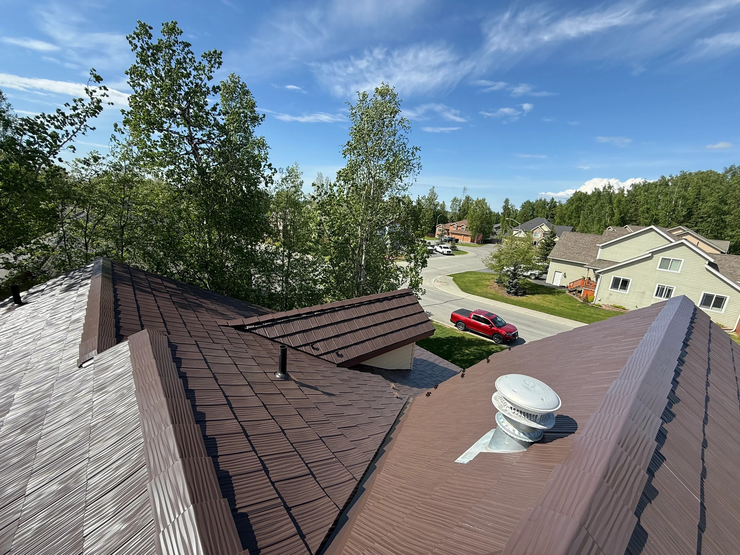 A modern single-family home with a brown metal roof and a red car in the driveway, surrounded by lush greenery and a blue sky.