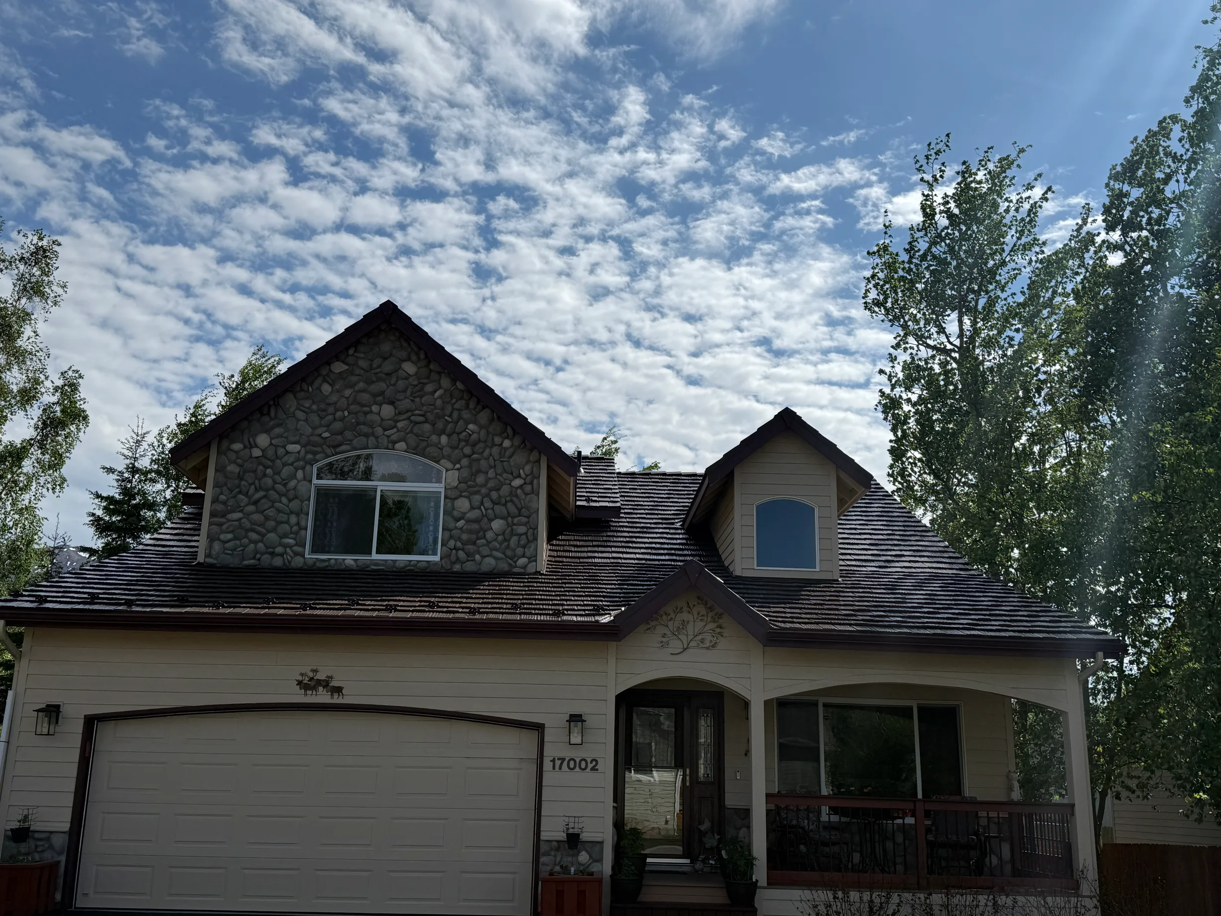 A cream and brown craftsman style house with a brown metal roof and a brown garage door, set against a lush green forested backdrop under a blue sky.