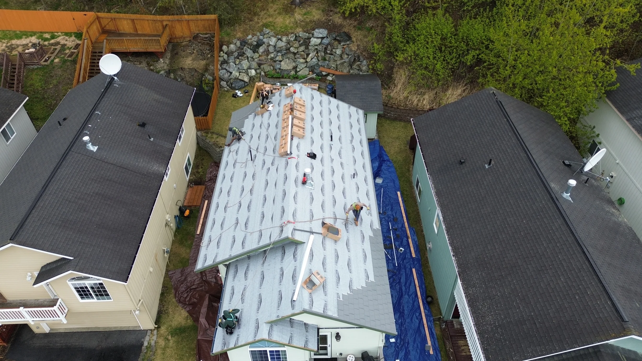 Aerial view of a ranch-style home with a gray metal roof being installed in a rural setting with lush greenery and blue skies