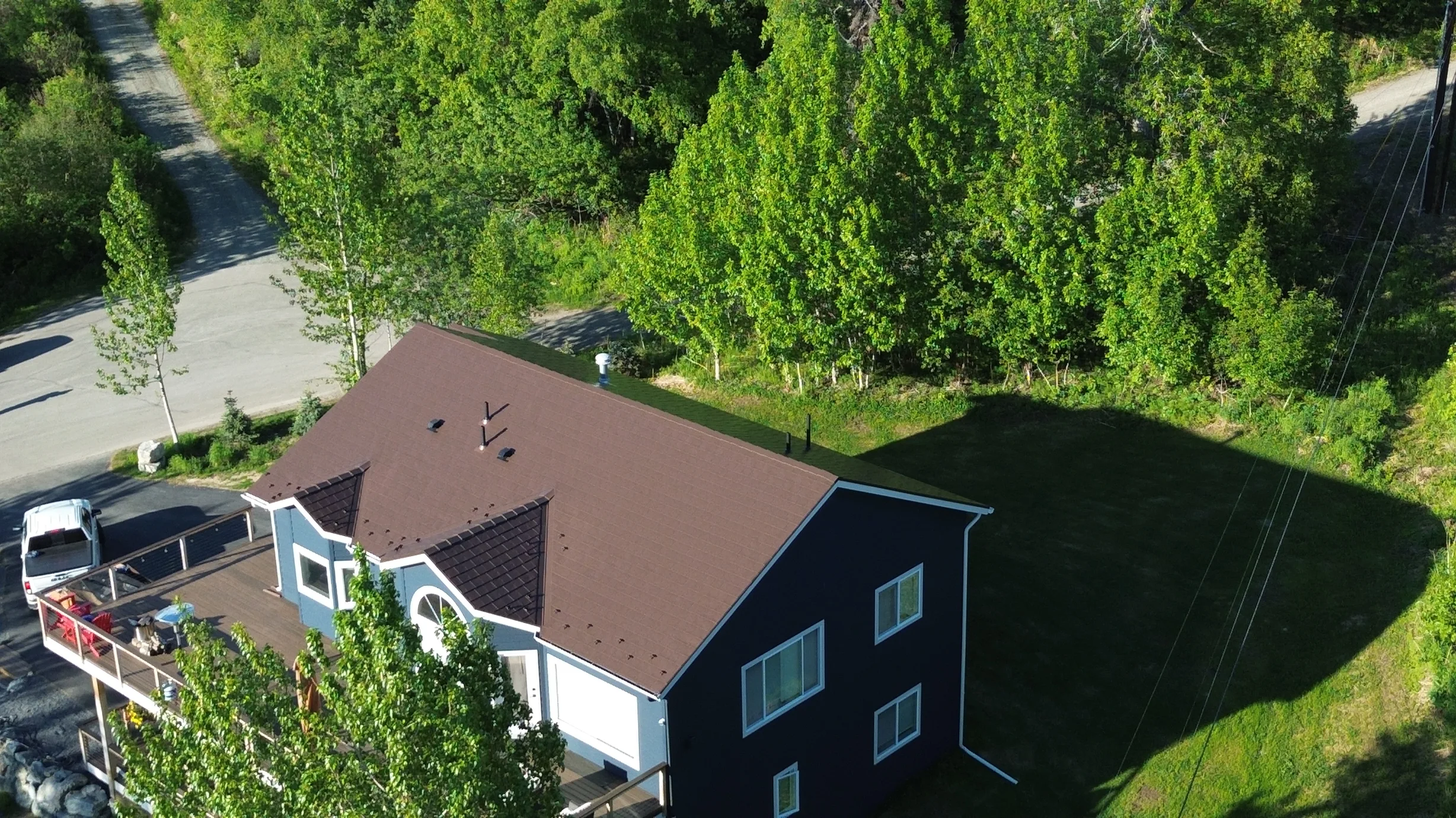 A two-story ranch-style home with a brown metal roof, white trim, and a brown deck, set against a lush green forest backdrop.