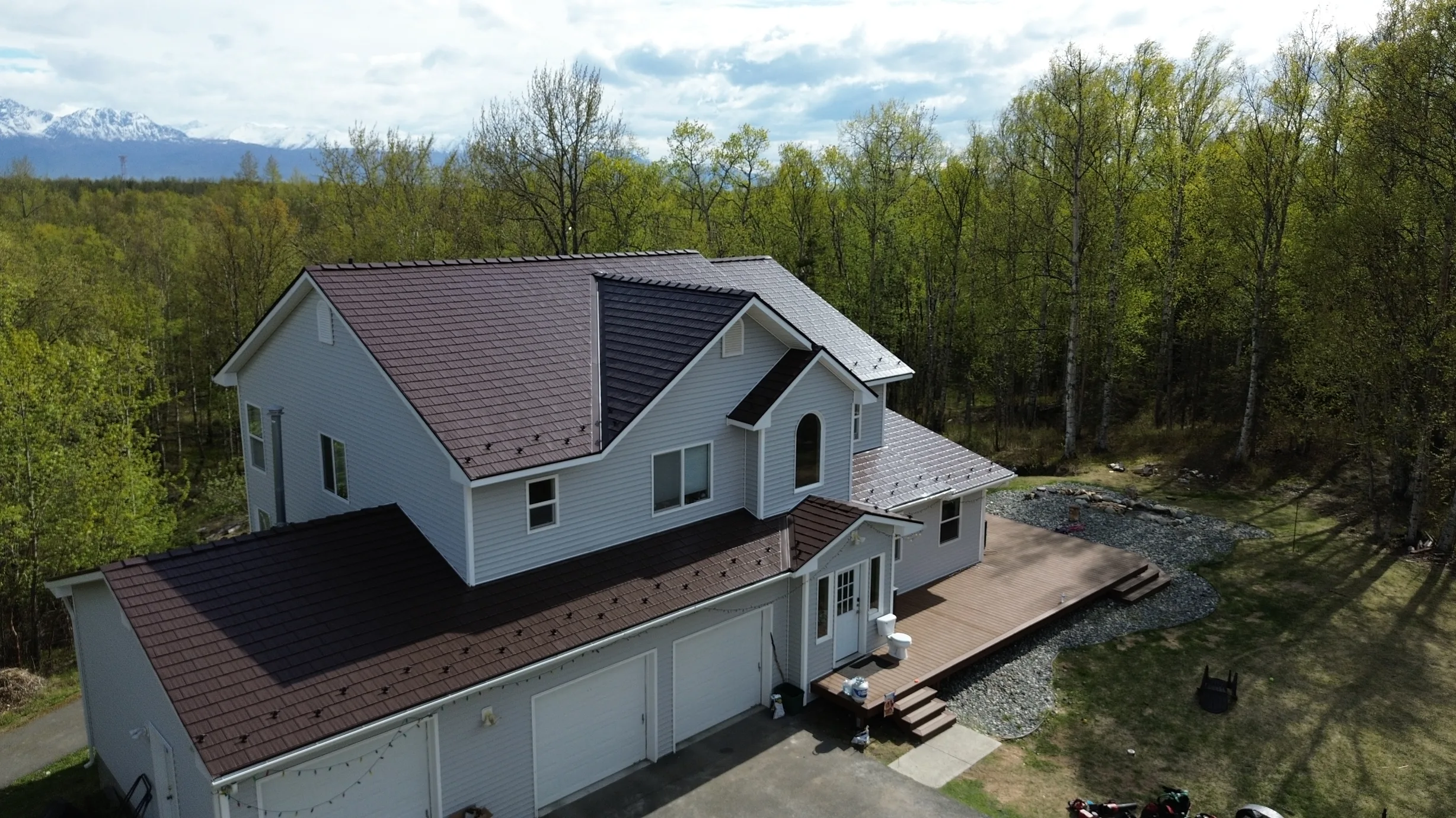 A white two-storey house with a brown metal roof in a wooded area.