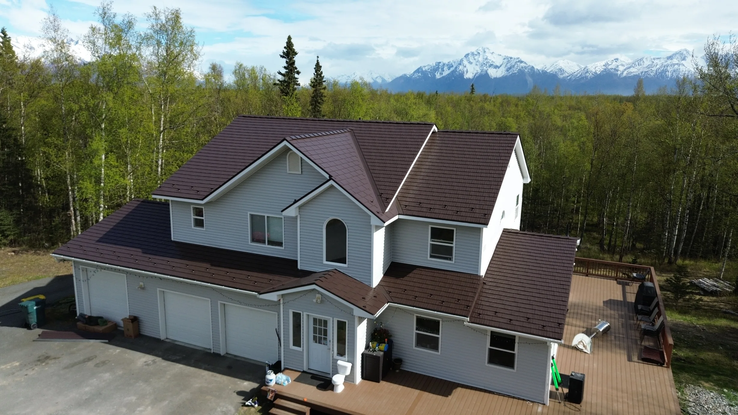 A two-story house with a brown metal roof and white siding, set amidst lush greenery and a mountain backdrop.