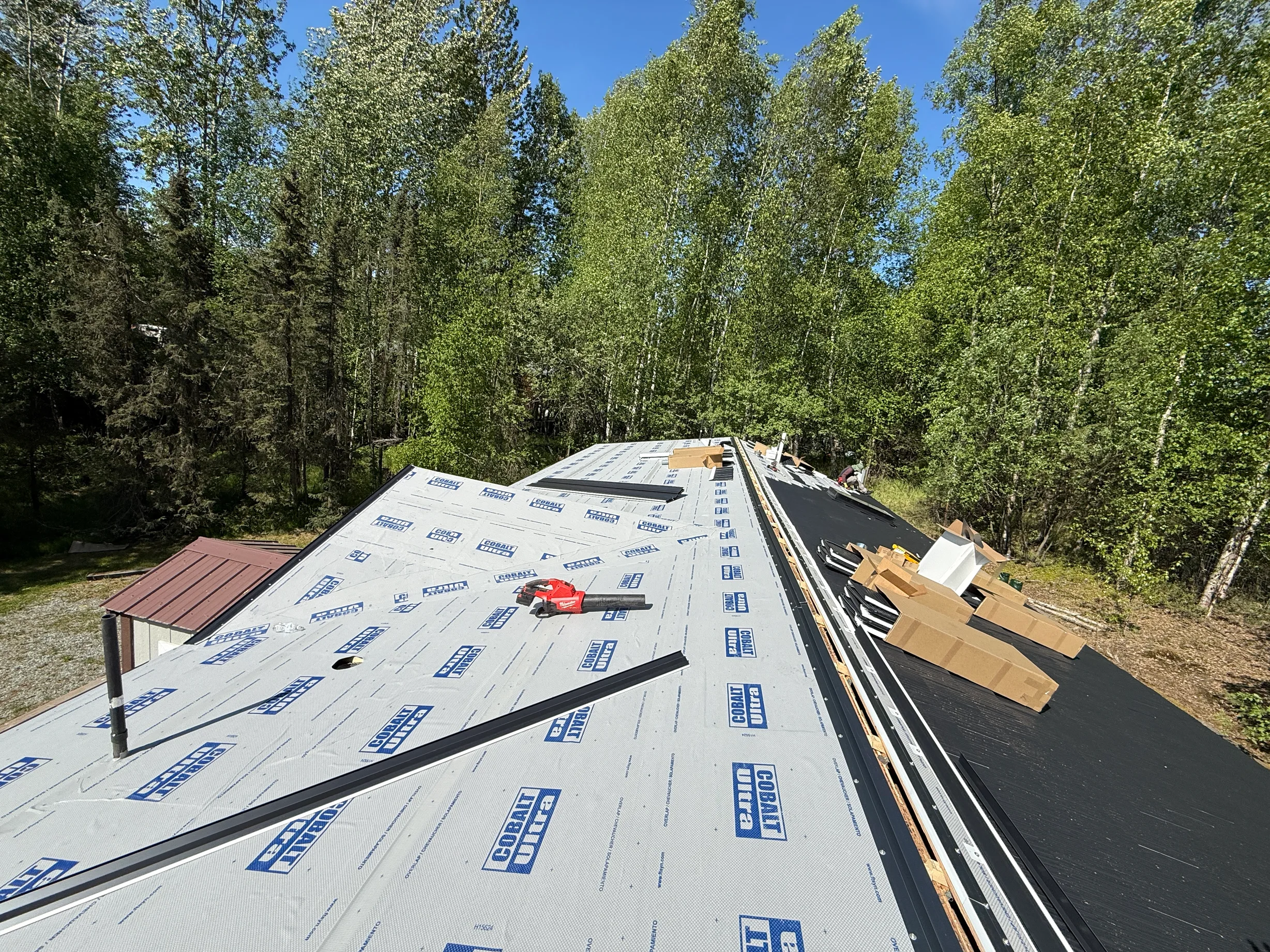 A metal roof under construction in a forested area with lush greenery and blue sky
